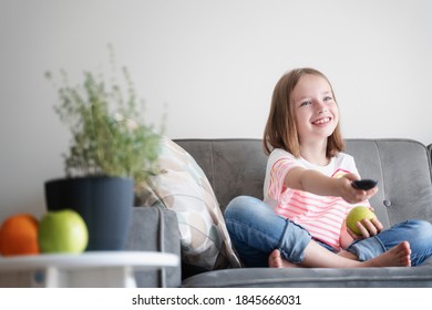 Adorable child girl 8 years old sits on a gray sofa with a green apple and a TV remote control in her hands
