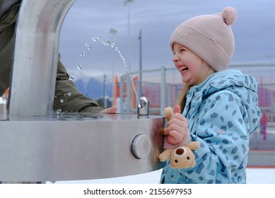 Adorable Child Girl Drinking From Outdoor Water Fountain And Smiles. Close Up. Kid Drink Tap Water In The Park In A Hat And Jacket