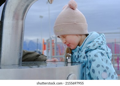 Adorable Child Girl Drinking From Outdoor Water Fountain And Smiles. Close Up. Kid Drink Tap Water In The Park In A Hat And Jacket