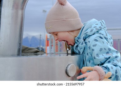 Adorable Child Girl Drinking From Outdoor Water Fountain And Smiles. Close Up. Kid Drink Tap Water In The Park In A Hat And Jacket