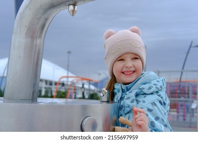 Adorable Child Girl Drinking From Outdoor Water Fountain And Smiles. Close Up. Kid Drink Tap Water In The Park In A Hat And Jacket