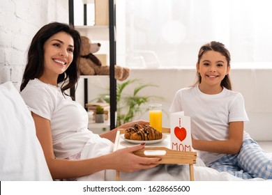 Adorable Child Sitting Near Mother Holding Tray With Breakfast, Mothers Day Card With Heart Sign And Mom Lettering