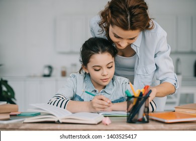 adorable child writing in copy book while doing schoolwork near mother