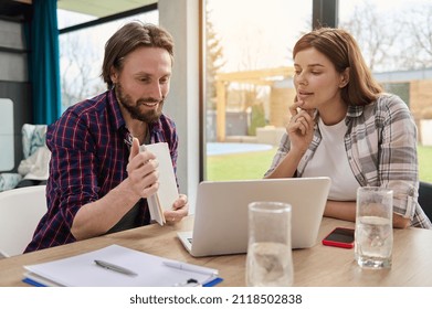 Adorable Couple Sitting At Home In Front Of Laptop, Showing To The Web Camera A Blank Page On Notepad While Having Video Call Against The Background Of Large Windows Overlooking The Garden In A Villa