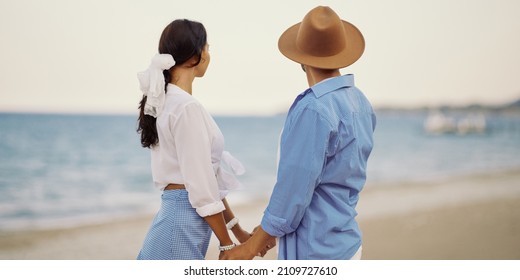 An Adorable Couple Of Young Adult Woman And Man In Summer Beach Outfits In Love Relaxing Outdoors Against The Beach In The Background. A Fashionable Romantic Couple Dating Outdoors