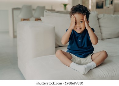 Adorable Curly Afro American Boy In Casual Wear Sitting On Comfy Big White Couch And Ready To Play, Stopping Counting And Uncovering His Eyes, About To Go Searching In Game Of Hide And Seek