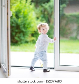 Adorable curly baby girl standing at a big glass door to the garden