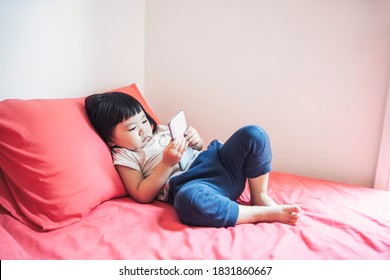 Adorable Cute Asian Little Kid Girl Addiction Addictive To Smartphone Mobile Cellphone Playing Device Tired Watching Video At Home In Bedroom Enjoying Having Fun Modern Childhood Technology Lifestyle