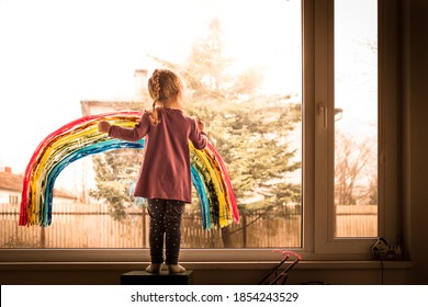Adorable, cute, beautiful, happy preschool kid girl painting colorful rainbow on window, concept of joy and hope during pandemic and social distancing