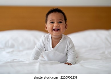 Adorable Cute Black Baby Lying On Tummy While Relaxing On Bed At Home, Closeup Portrait Of Smiling Little Infant African American Boy Or Girl Resting On White Linens In Bedroom, Copy Space