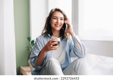 Adorable cute funny woman with a mug of black tea talking on the phone sitting on the windowsill in the kitchen, having fun, and enjoying pleasant conversation with her sister or best friend