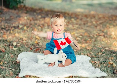 Adorable Cute Little Caucasian Baby Toddler Girl Waving Canadian Flag In Park Outdoors. Kid Child Citizen Sitting On Ground In Park And Celebrating Canada Day On 1st Of July.