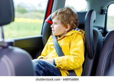 Adorable Cute Preschool Kid Boy Sitting In Car In Yellow Rain Coat. Little School Child In Safety Car Seat With Belt Enjoying Trip And Journey. Safe Travel With Kids And Traffic Laws Concept.