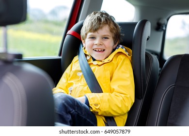 Adorable Cute Preschool Kid Boy Sitting In Car In Yellow Rain Coat. Little School Child In Safety Car Seat With Belt Enjoying Trip And Journey. Safe Travel With Kids And Traffic Laws Concept.