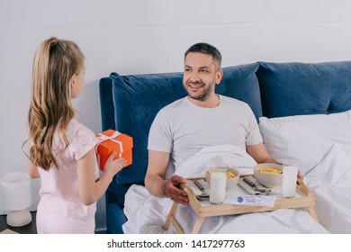 Adorable Daughter Holding Fathers Day Gift Box While Standing Near Father Having Breakfast In Bed