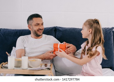 Adorable Daughter Presenting Fathers Day Gift Box To Smiling Dad Having Breakfast In Bed