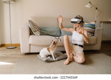 Adorable Dog Watches In Bewilderment As Young Slender Blonde Woman Using Virtual Reality Simulator Sitting On Floor With Hands Raised In Surprise. VR Glasses Headset  Watching 360 Degree Video