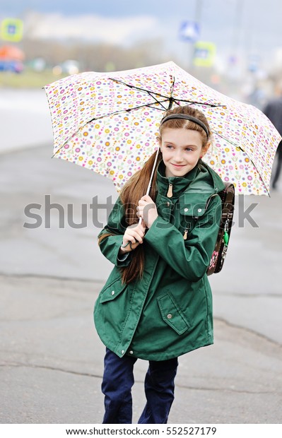Adorable, elegant school\
aged kid girl ,holding colorful umbrella walking in the city street\
in rainy day