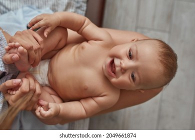 Adorable Emotional Baby Laughing Lying Naked On Female Knees. Unrecognised Female Holding Baby On Knees, Touching Its Barefeet. Positive Baby Enjoying Tactile Contact With Mother.