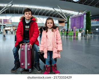 Adorable European Children In Warm Clothes Sit On Suitcases And Look At The Camera In The Departure Area Of The International Airport. Family Holidays,winter Tourism And Air Travel Concept. Copy Space