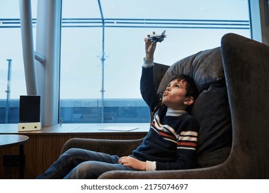 Adorable European Teenage Boy Plays With Toy Airplane Sitting By Panoramic Windows Overlooking The Runway At Sunset While Waiting To Board Flight At International Airport Departure Terminal