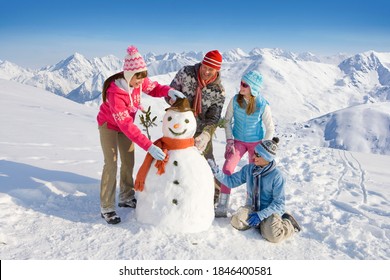 An Adorable Family Having Fun While Building And Decorating A Snowman Together In Front Of A Magnificent Range Of Snow Mountains