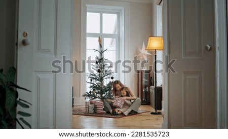 Adorable family unpacking gifts near xmas tree. Positive mother and curious daughter looking at present at cozy interior. Tender woman embracing child at Christmas decorated home. New Year concept