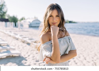 Adorable fantastic woman with style hairstyle and big eyes smiles to camera with great emotions. Young effective woman wearing summer dress with bare shoulders on background on the sunny summer beach