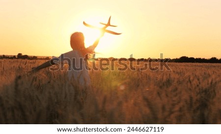 Adorable female kid silhouette running with aircraft toy at cinematic sunset sunrise wheat field back view. Happy little girl child playing pilot game with plane plaything enjoy childhood at dusk