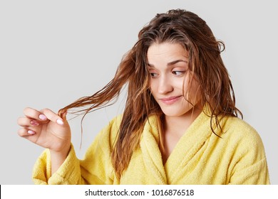 Adorable Female Looks At Split Ends After Taking Shower, Has Damaged Hair, Wears Domestic Clothes, Isolated Over White Background. Frustarted Woman Dissatisfied With Dry Frizzy Hair