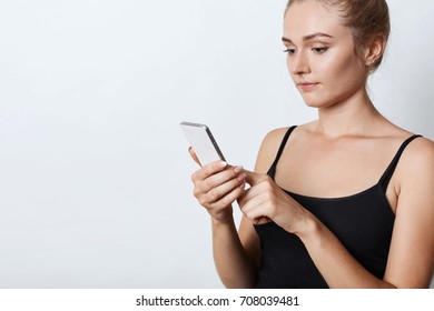 Adorable Female With Tied Hair, Wearing Black Casual Clothes, Holding Modern Mobile Phone In Hand, Reading Attentively Electronic Book. Female Student Preparing For Final Exams Using Smart Phone