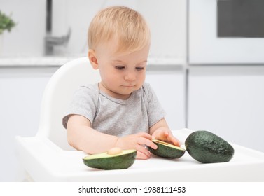 Adorable Funny Caucasian Blonde Baby Girl Eating Green Avocado At Highchair In Kitchen. Kid Supplementary Healthy Food. Fresh Vegetables For Child .Selective Focus