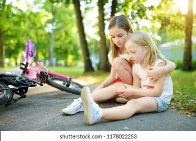 Adorable Girl Comforting Her Little Sister After She Fell Off Her Bike At Summer Park. Child Getting Hurt While Riding A Bicycle. Active Family Leisure With Kids.