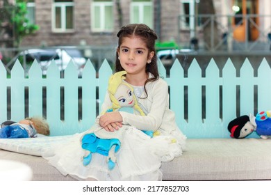 Adorable Girl Hugging Her Handmade Soft Toy And Sitting On The Floor In Kindergarten With Multi Colored Interior