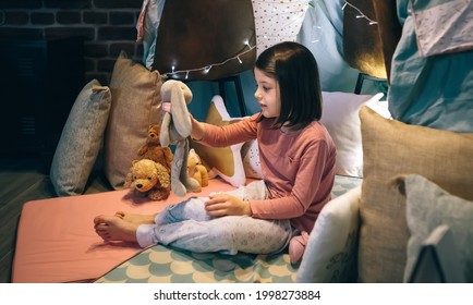 Adorable Girl In Pajamas Playing Alone With Teddies In A Play Shelter Made Of Bed Sheets And Chairs
