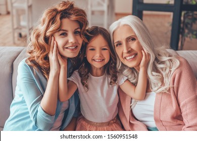 adorable girl sitting between mother and grandmother and touching their faces with hands sitting on the couch