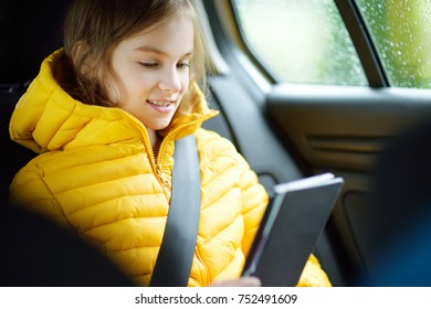 Adorable girl sitting in a car and reading her ebook on rainy autumn day. Child entertaining herserf on a road trip. Traveling by car with kids.