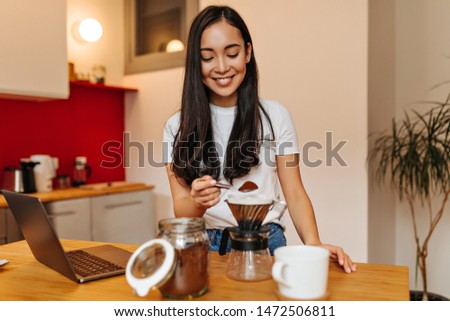 Adorable girl with smile makes coffee in kitchen and looks into laptop