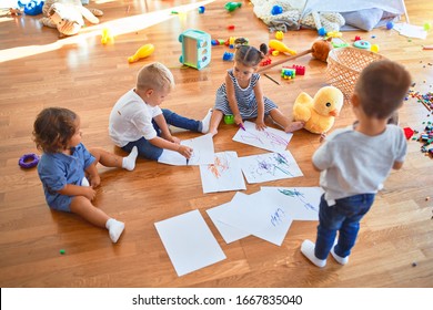 Adorable group of toddlers sitting on the floor drawing using paper and pencil around lots of toys at kindergarten