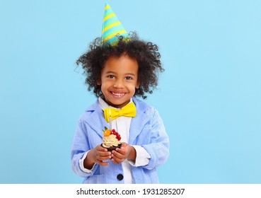 Adorable happy African American little boy in bow tie and party hat smiling and looking at camera while standing against blue background with yummy birthday cupcake in hands