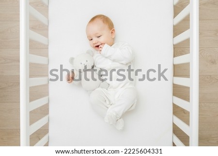 Adorable happy baby boy in white bodysuit hugging teddy bear and  lying down on back on mattress in crib at home room. Top view. 6 months old infant playing with first friend. Closeup. 