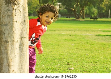 Adorable Happy Kid With Curly Hair Peeking Around The Tree Playing Hide And Seek In A Park At Summer Evening