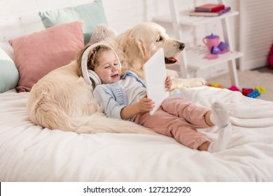 Adorable Happy Kid Listening Music With Tablet And Leaning On Golden Retriever Dog On Bed In Children Room