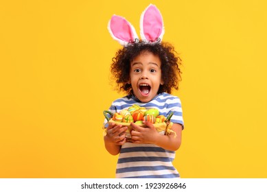 Adorable happy little African American boy with curly hair and bunny ears on head screaming with astonishment  while holding Easter basket while standing against yellow background  - Powered by Shutterstock