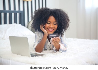 Adorable Hipster Dark-skinned Woman With Afro Hairstyle Checking Her News Feed Or Messaging Via Social Networks, Using Free Wi-fi On Mobile Phone, Smiling, Sitting At Home In Front Of Laptop Computer