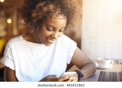 Adorable hipster dark-skinned woman with Afro hairstyle checking her news feed or messaging via social networks, using free wi-fi on mobile phone, smiling, sitting at cafe in front of laptop computer - Powered by Shutterstock