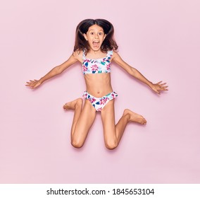 Adorable hispanic child girl on vacation wearing bikini surprised with open mouth. Jumping over isolated pink background