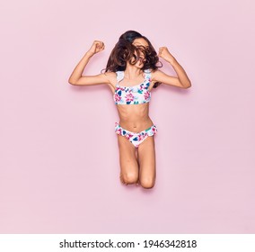 Adorable Hispanic Child Girl On Vacation Wearing Bikini Surprised With Open Mouth. Jumping Over Isolated Pink Background