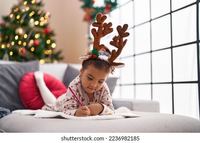 Adorable hispanic girl drawing on notebook lying on sofa by christmas tree at home - Powered by Shutterstock