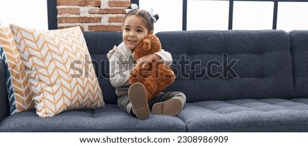 Adorable hispanic girl hugging teddy bear sitting on sofa at home